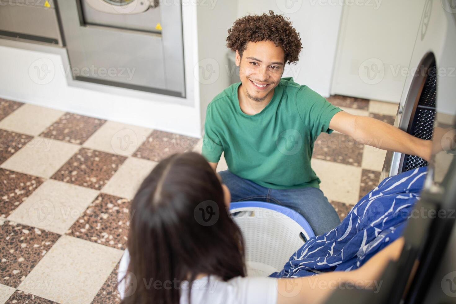 A young man smiles at a woman as they work together in a laundromat, loading clothes into a washing machine. The bright space features modern appliances and tiled flooring. photo