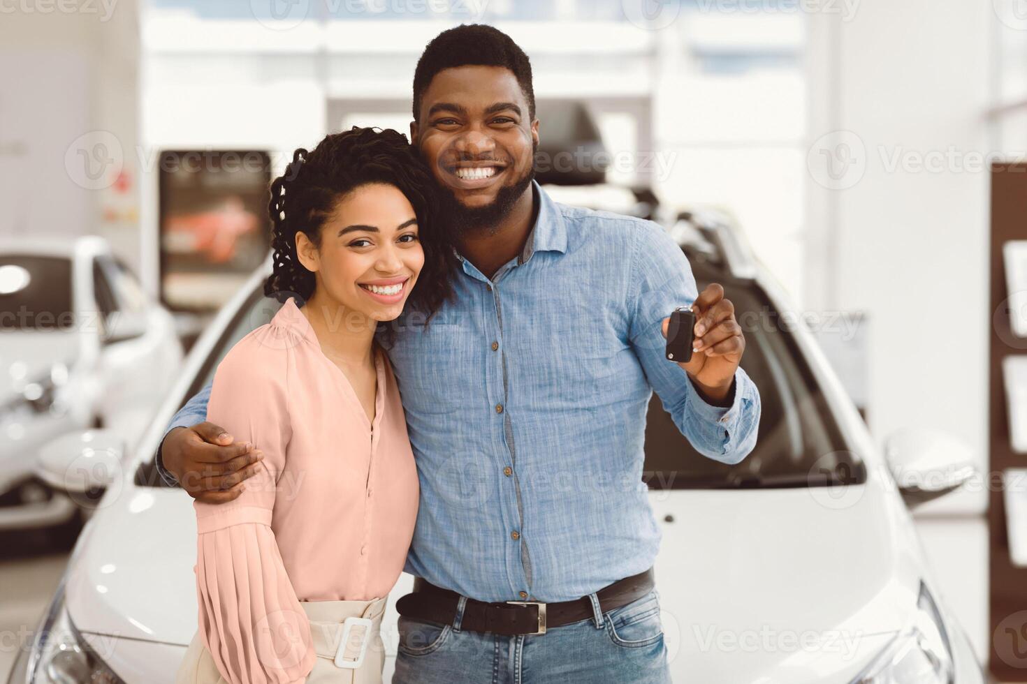 African American Couple Holding New Automobile Key Embracing Standing Near New Family Car In Dealership Center. photo