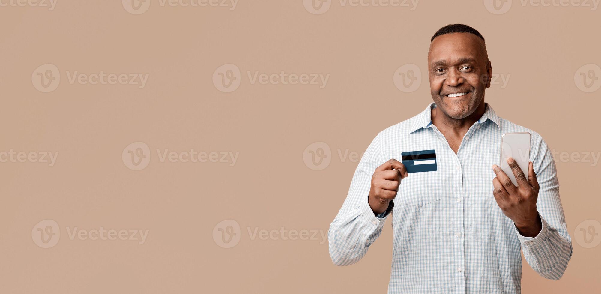 Online banking concept. Cheerful african american man checking his card balance with mobile application, orange studio background photo