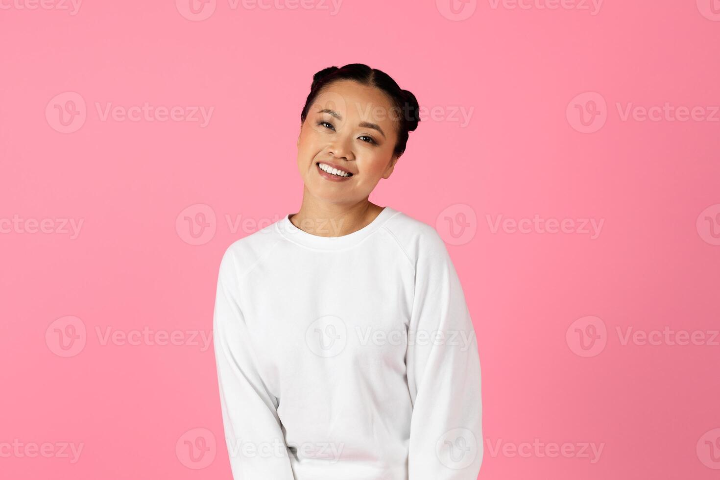 A cheerful young Asian woman stands confidently with a bright smile, wearing a simple white sweater. The vibrant pink backdrop enhances her joyful expression, creating a lively atmosphere. photo