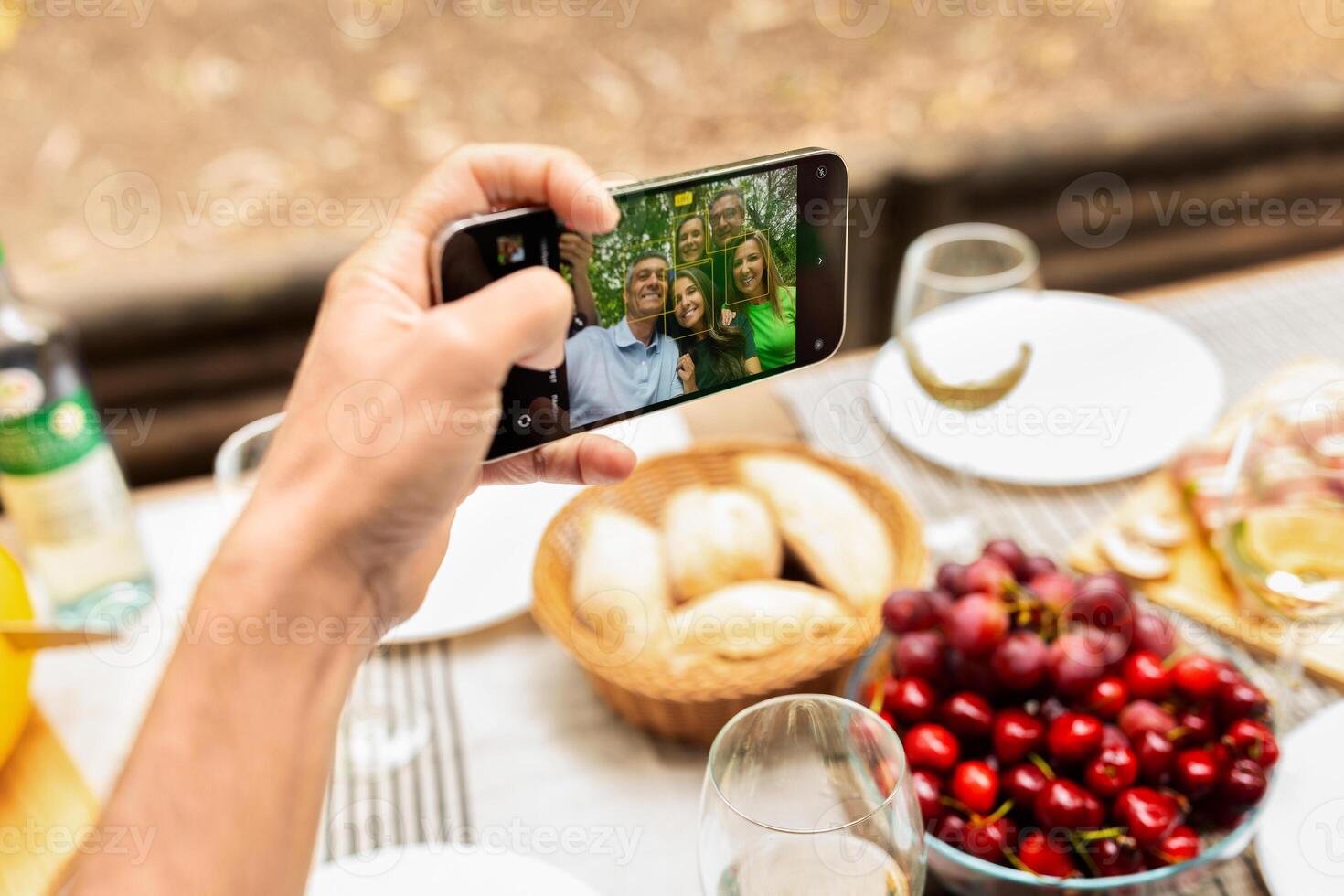A hand holds a smartphone capturing a group selfie during a family gathering outdoors. Delicious food items like fruits and bread are displayed on the table, enhancing the cheerful atmosphere. photo