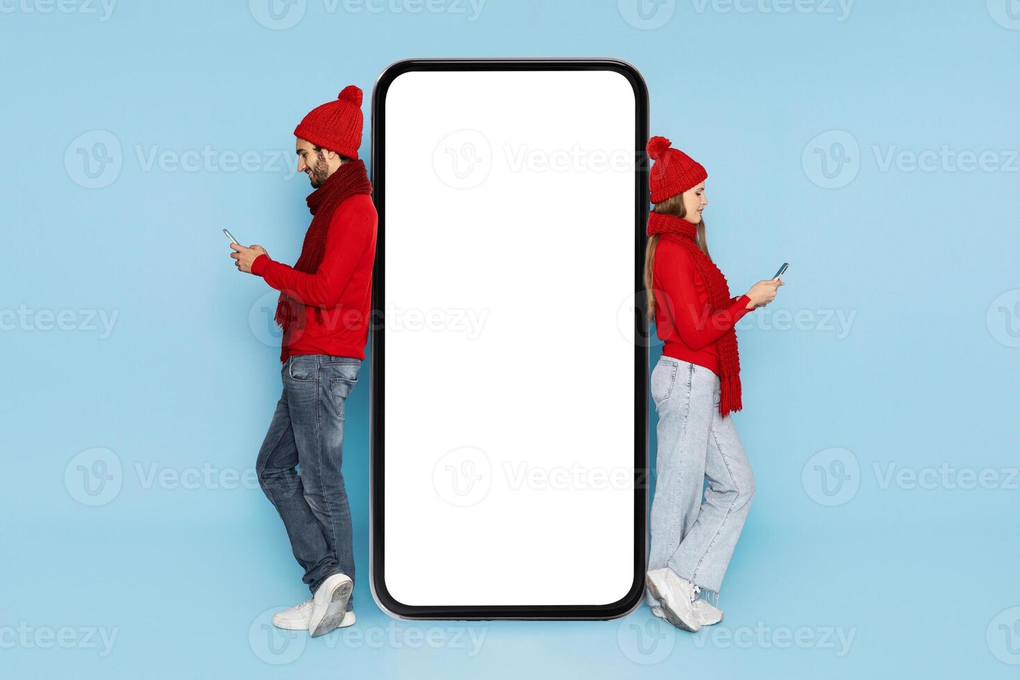 A young couple stands back to back wearing red sweaters and hats, focused on their smartphones. They are positioned next to a large blank screen, suggesting a playful or social setting. photo