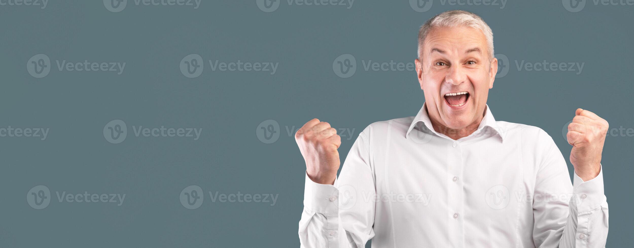 Yes. Excited senior man screaming with joy, raising clenched fists in the air isolated over blue studio wall, panorama photo