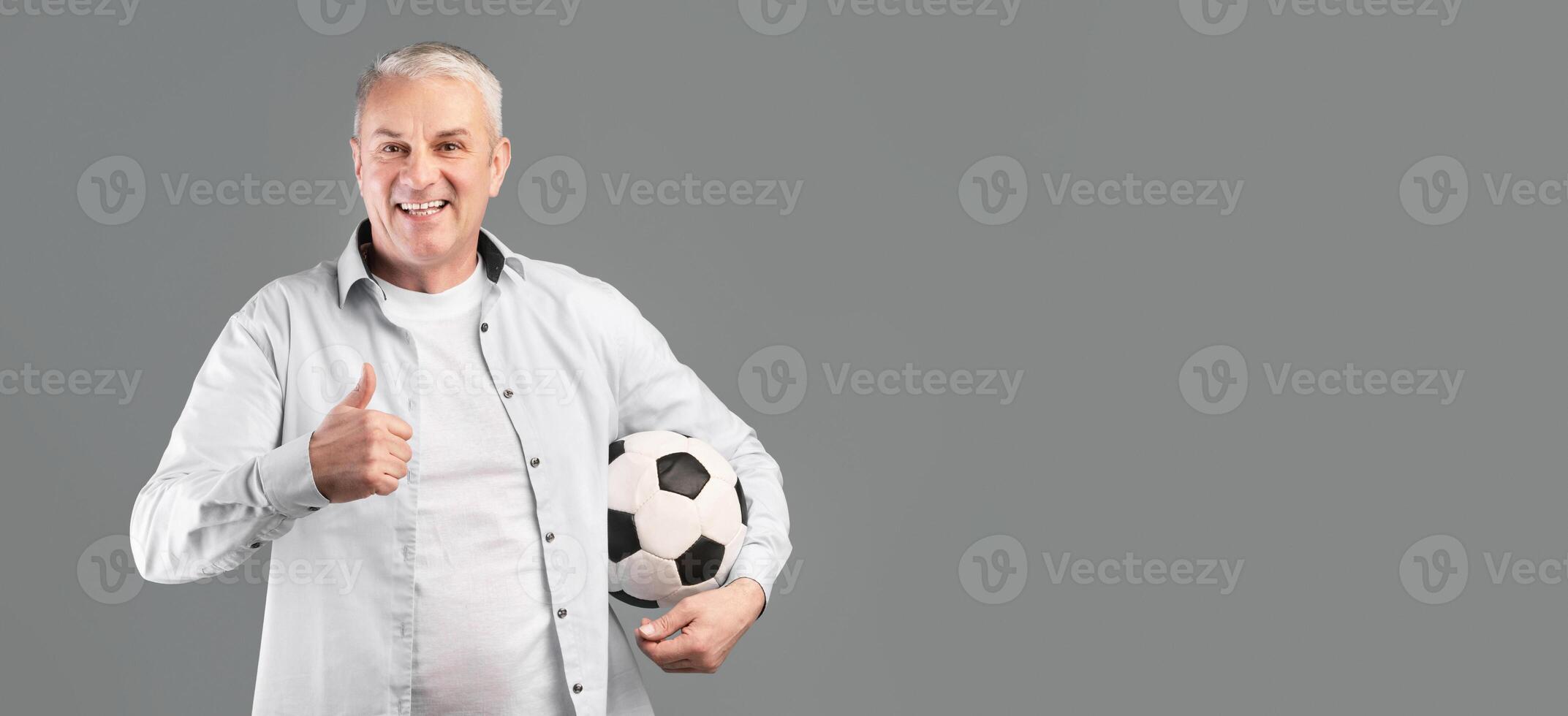 Mature Football Fan. Glad senior man posing with soccer ball, showing thumbs up gesture, gray studio wall photo