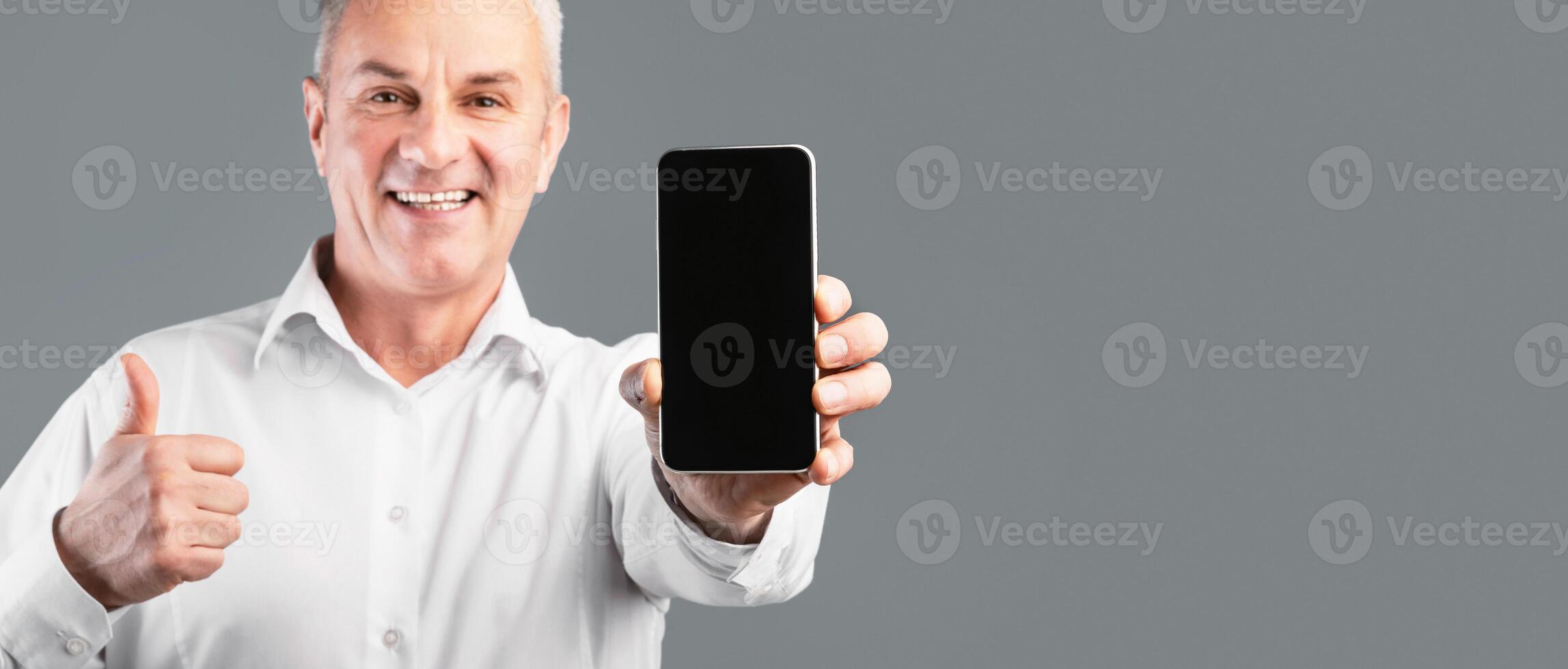 Useful App. Middle-aged man holding smartphone with blank black screen, showing thumbs up, gray studio wall, mock up photo