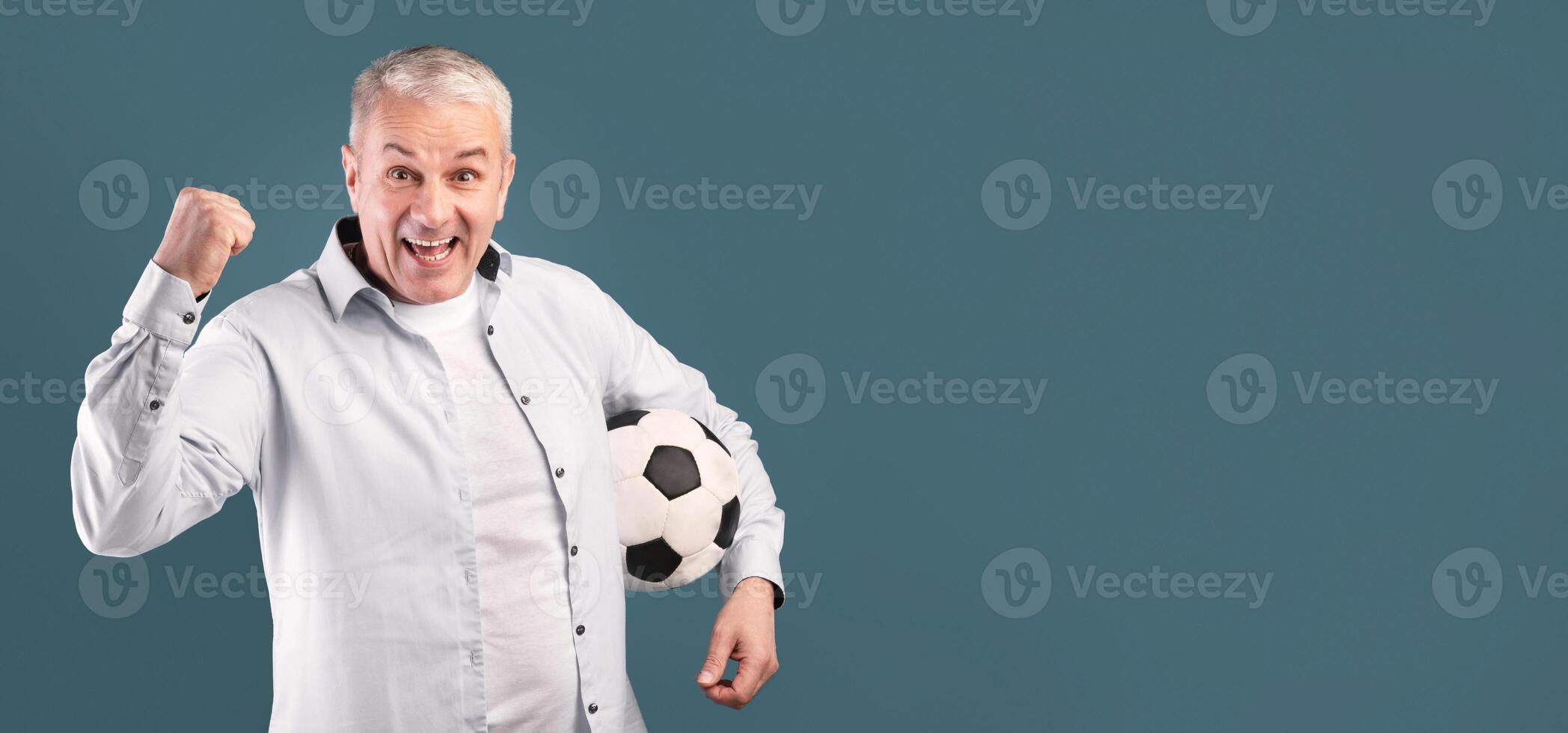 Go On. Excited middle-aged man making winner gesture with soccer ball on blue studio wall, free space photo