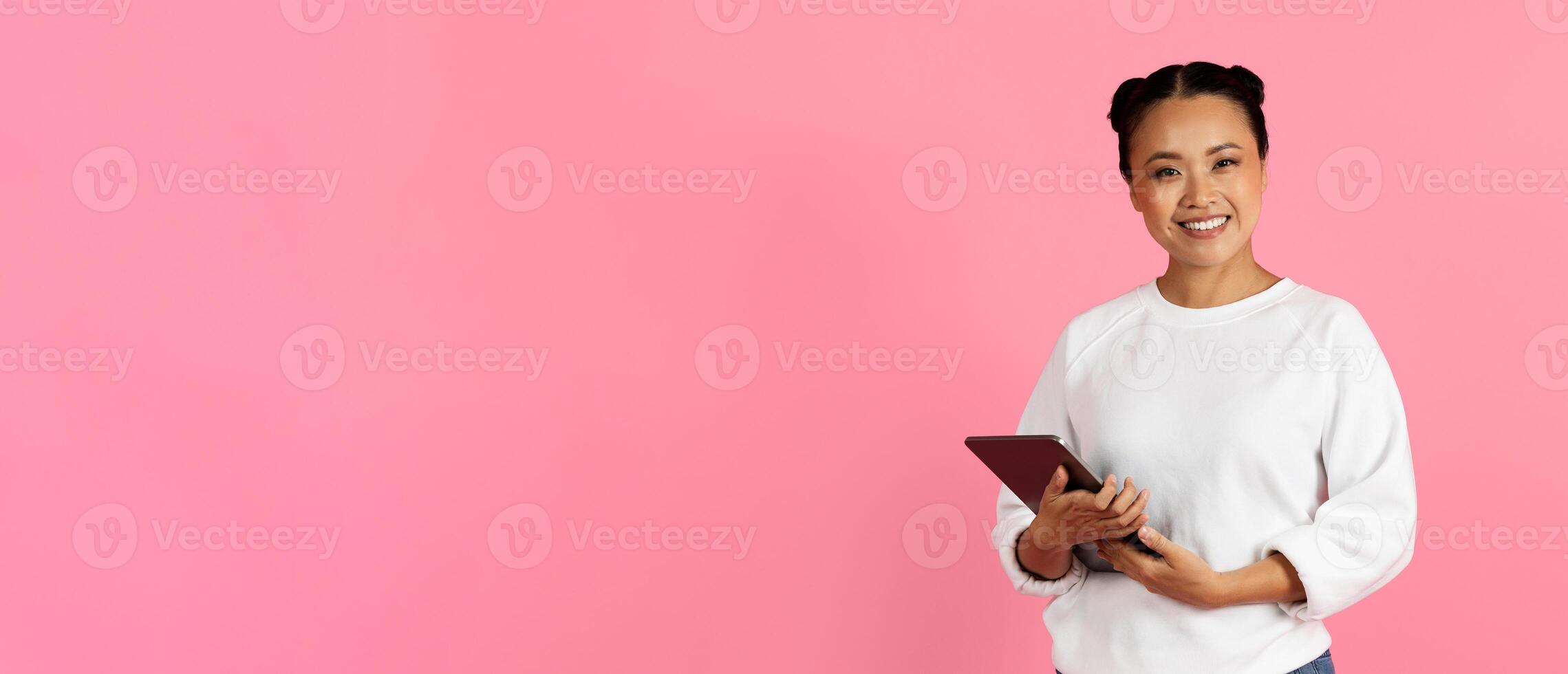 A cheerful young woman stands confidently, holding a tablet and smiling warmly. She is dressed casually against a vibrant pink backdrop, suggesting positivity and creativity, copy space photo