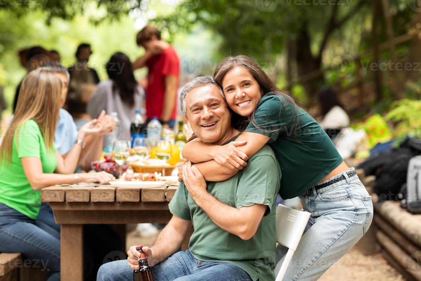 A joyful family gathering is happening in a park. A man is hugging a young woman, both smiling, surrounded by loved ones enjoying food and drinks on a sunny day. photo
