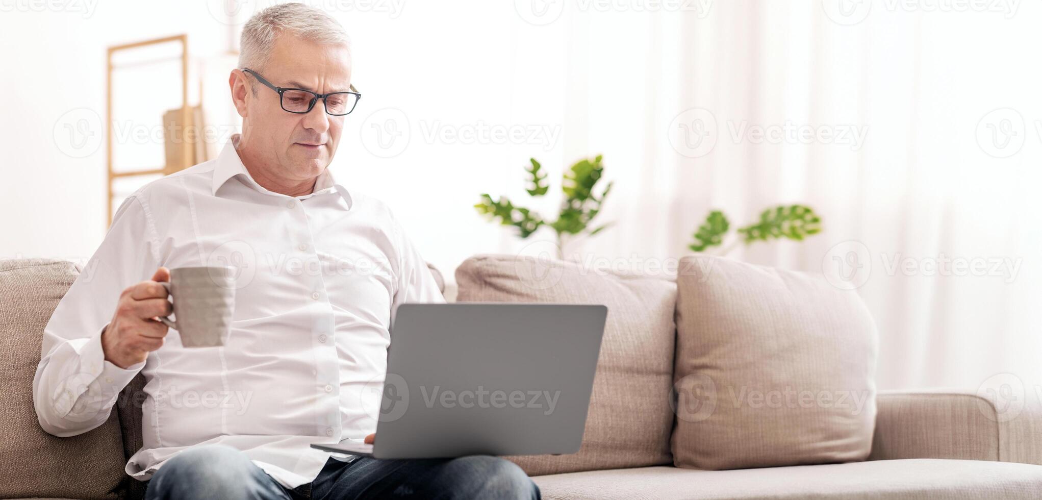 Serious Mature Man Holding Laptop Computer And Coffee Mug, Sitting On Sofa At Home, Empty Space photo