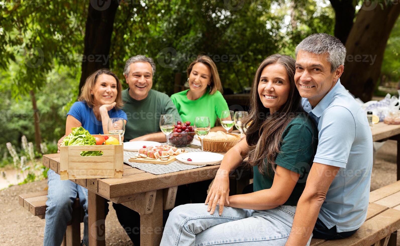 A group of five friends gathers around a picnic table in a verdant park, sharing food and laughter on a warm afternoon, with fruits and snacks spread out before them. photo