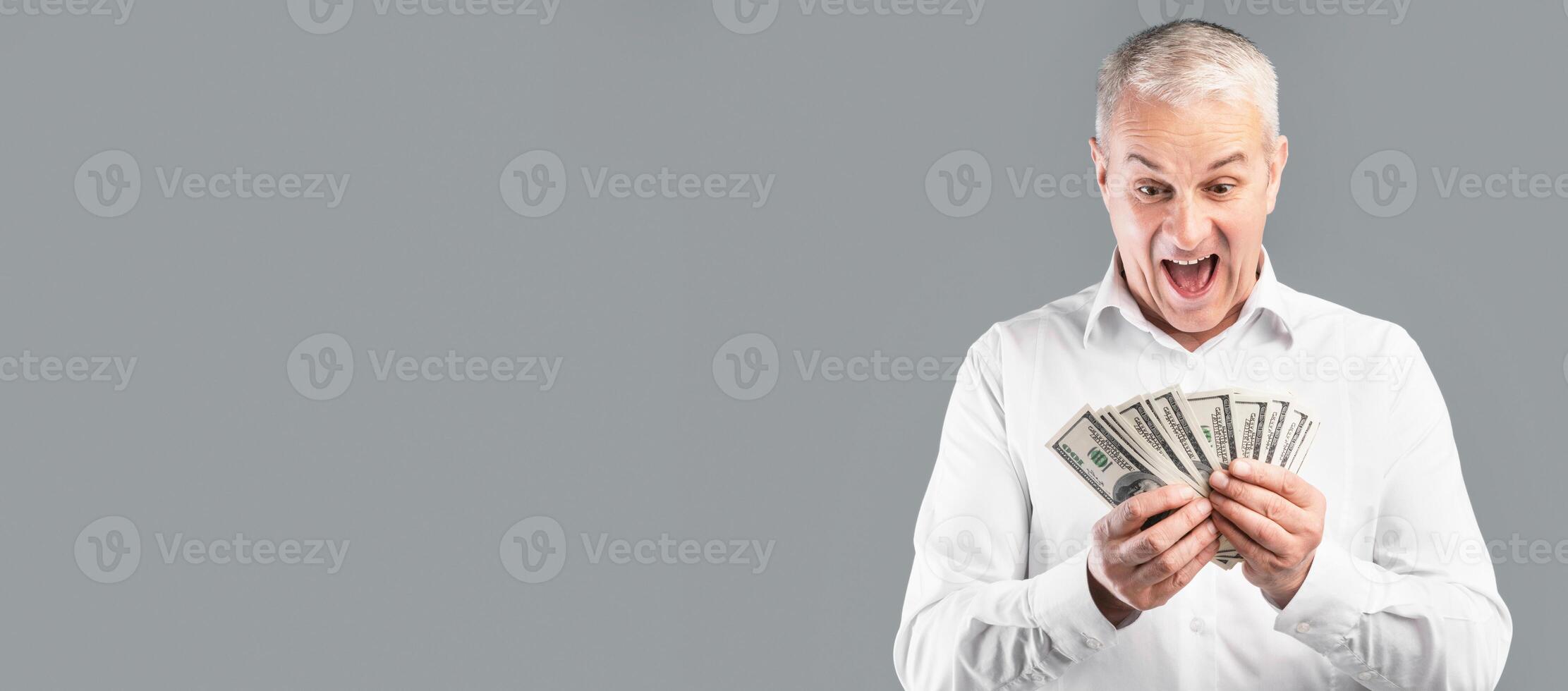 Big Win. Excited Senior Man Posing With Bunch Of Money, Holding Dollar Banknotes, Gray Studio Wall, Empty Space photo