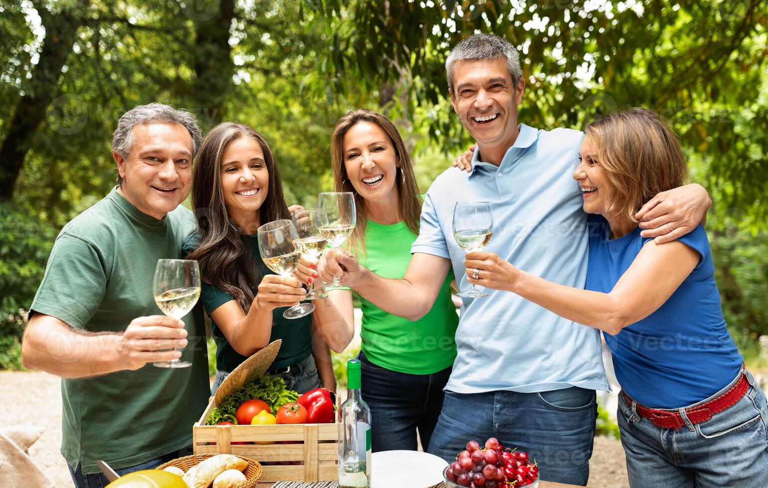 A cheerful gathering of five friends toasting with glasses of wine at a picnic table filled with fresh produce, surrounded by lush trees on a sunny day. photo