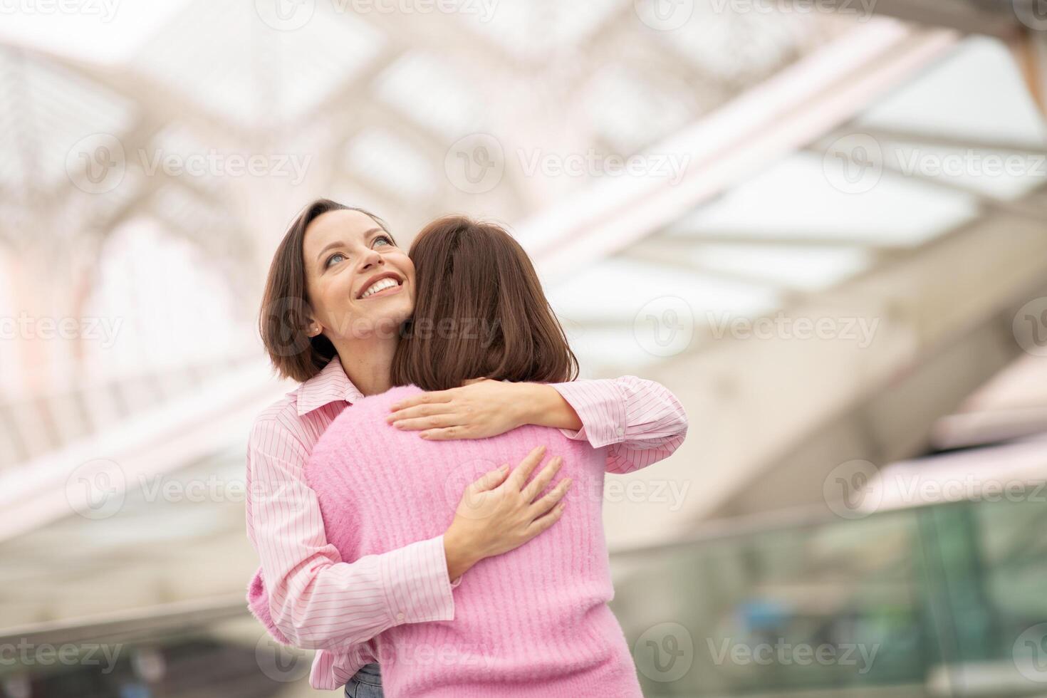 Two women smile and hug each other warmly in a contemporary city environment, showcasing friendship and connection under a unique architectural structure. photo