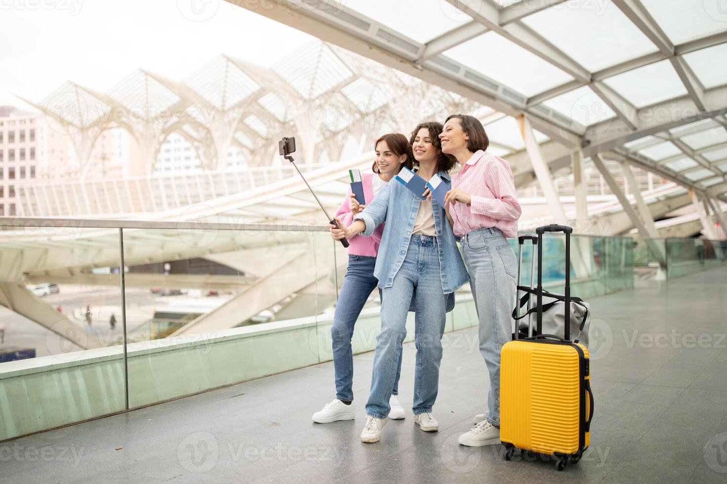 Three friends pose together for a selfie at a contemporary train station. The yellow suitcase rests nearby as they enjoy their trip and capture a memorable moment. photo