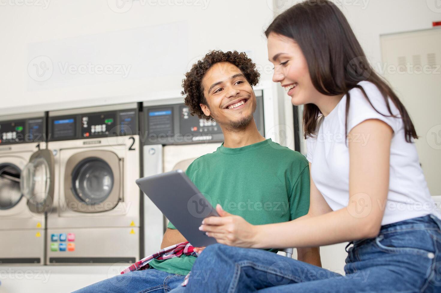 Two young adults sit side by side in a laundromat, smiling and engaging with a tablet as they wait for their laundry to finish in the background. photo