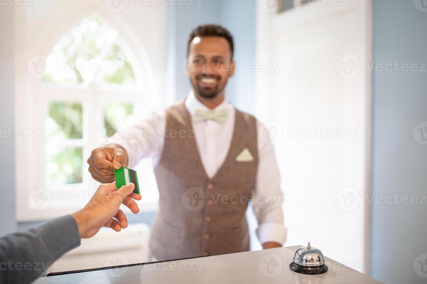 A man with a bow tie is handing a credit card to a person at a hotel reception desk. A bell is in the foreground on the counter. photo