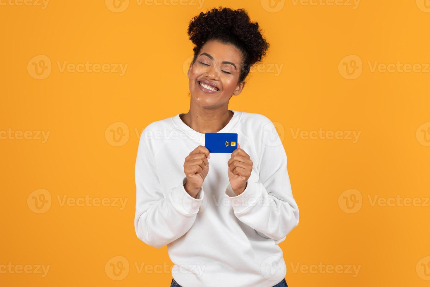 A smiling young woman wearing a white sweater confidently displays a blue card in front of a vibrant orange backdrop, radiating happiness and enthusiasm. photo