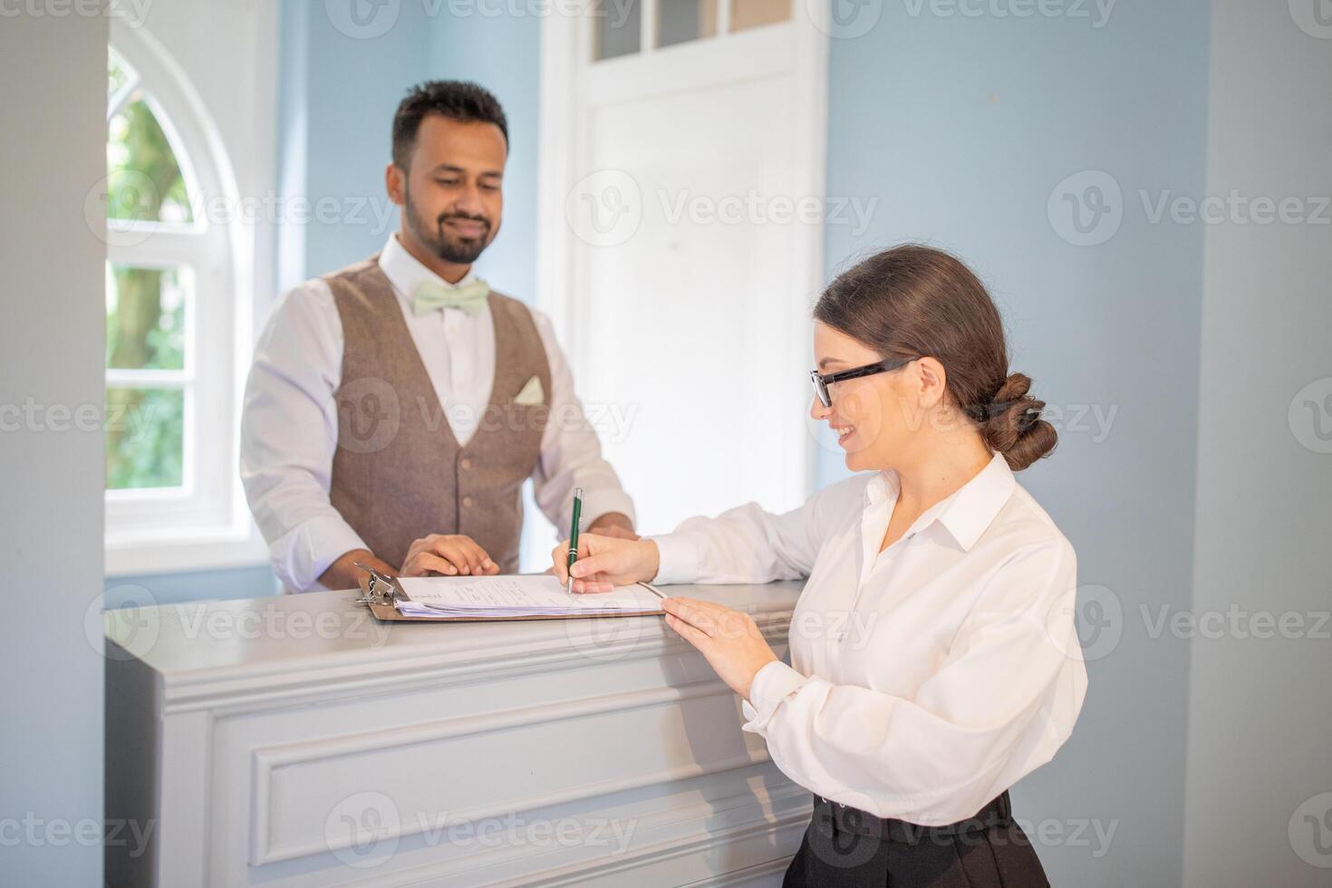 A receptionist is helping a guest with check-in at a stylish hotel lobby. The warm atmosphere and attentive service create a welcoming experience for visitors. photo