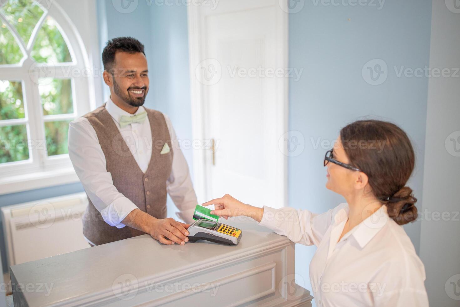 A smiling hotel clerk assists a guest during checkout in a well-lit reception area. The guest is making a payment with a card, creating a friendly interaction. photo