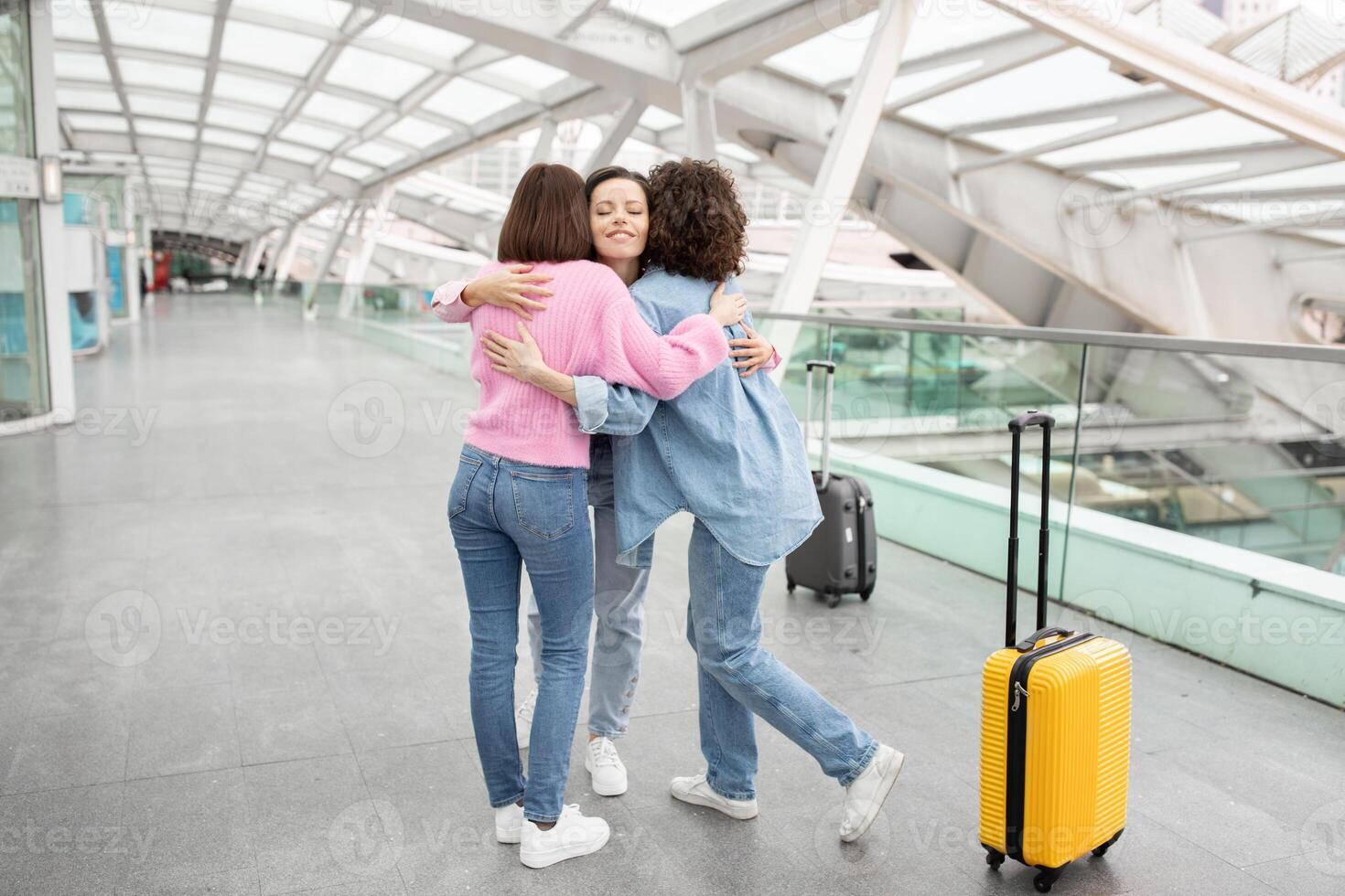 Three friends share a heartfelt hug at a bustling airport terminal. One is wearing a pink sweater, and the others are in a light blue outfit, with a yellow suitcase nearby, ready for their journey. photo