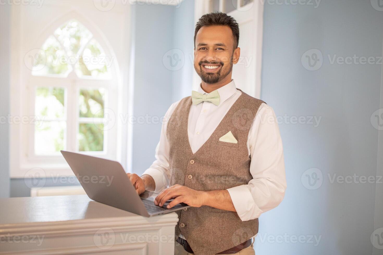 A man with a beard, wearing a white shirt, brown vest, and light green bow tie, receptionist smiles while looking at the camera. He is standing in front of a window with a white frame at hotel desk photo