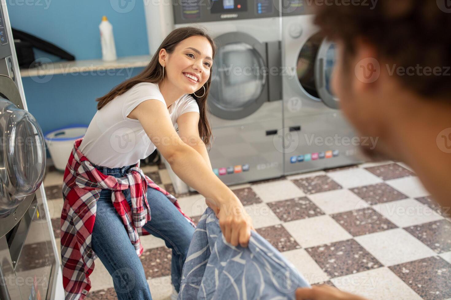 A cheerful young woman is folding laundry together with a friend in a colorful laundromat. The bright blue walls and modern appliances create a welcoming atmosphere. photo