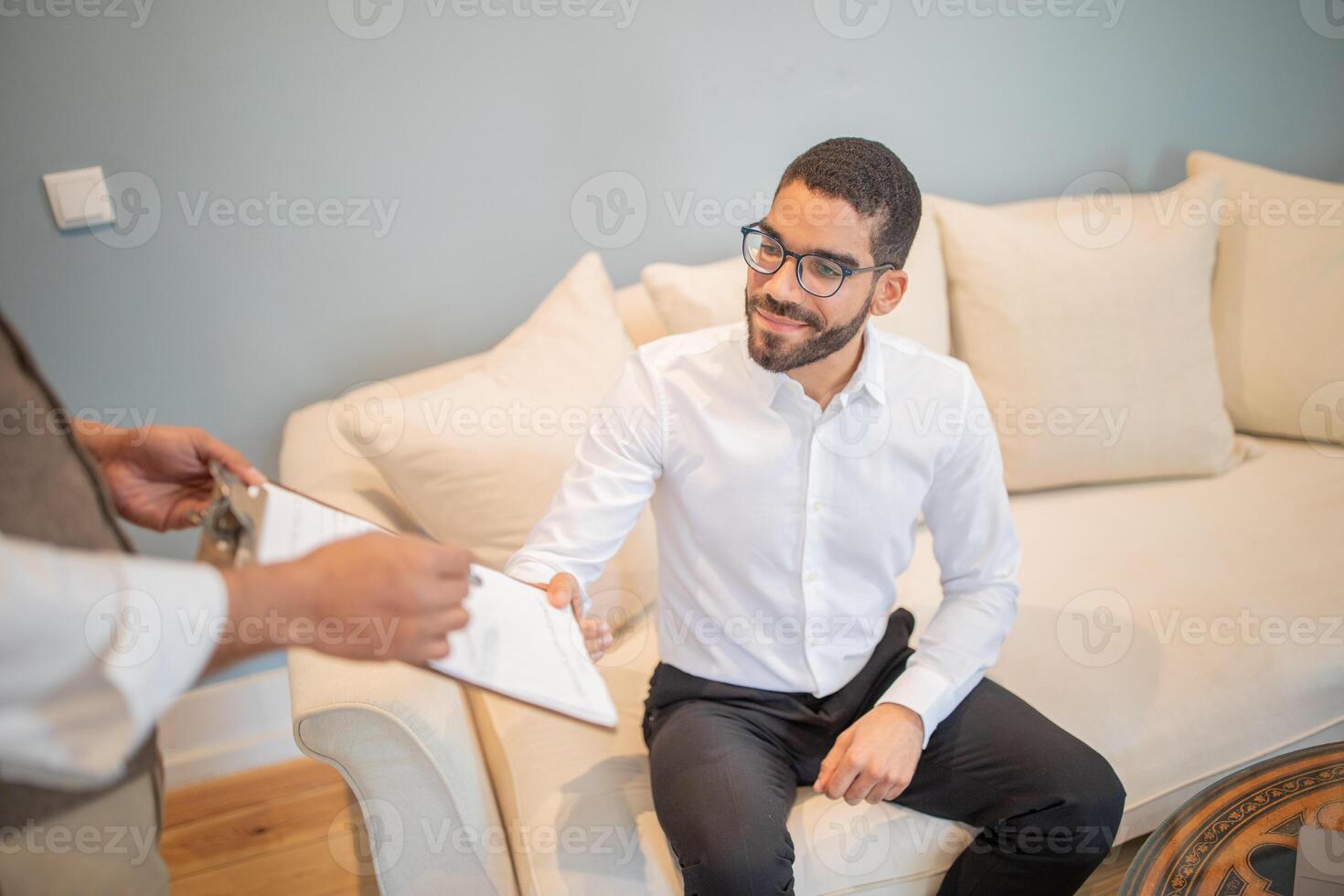 A man in a white shirt with a beard and glasses is sitting on a white couch. He is looking to the side, smiling. An unseen person is handing him a document, which he is accepting with his left hand. photo
