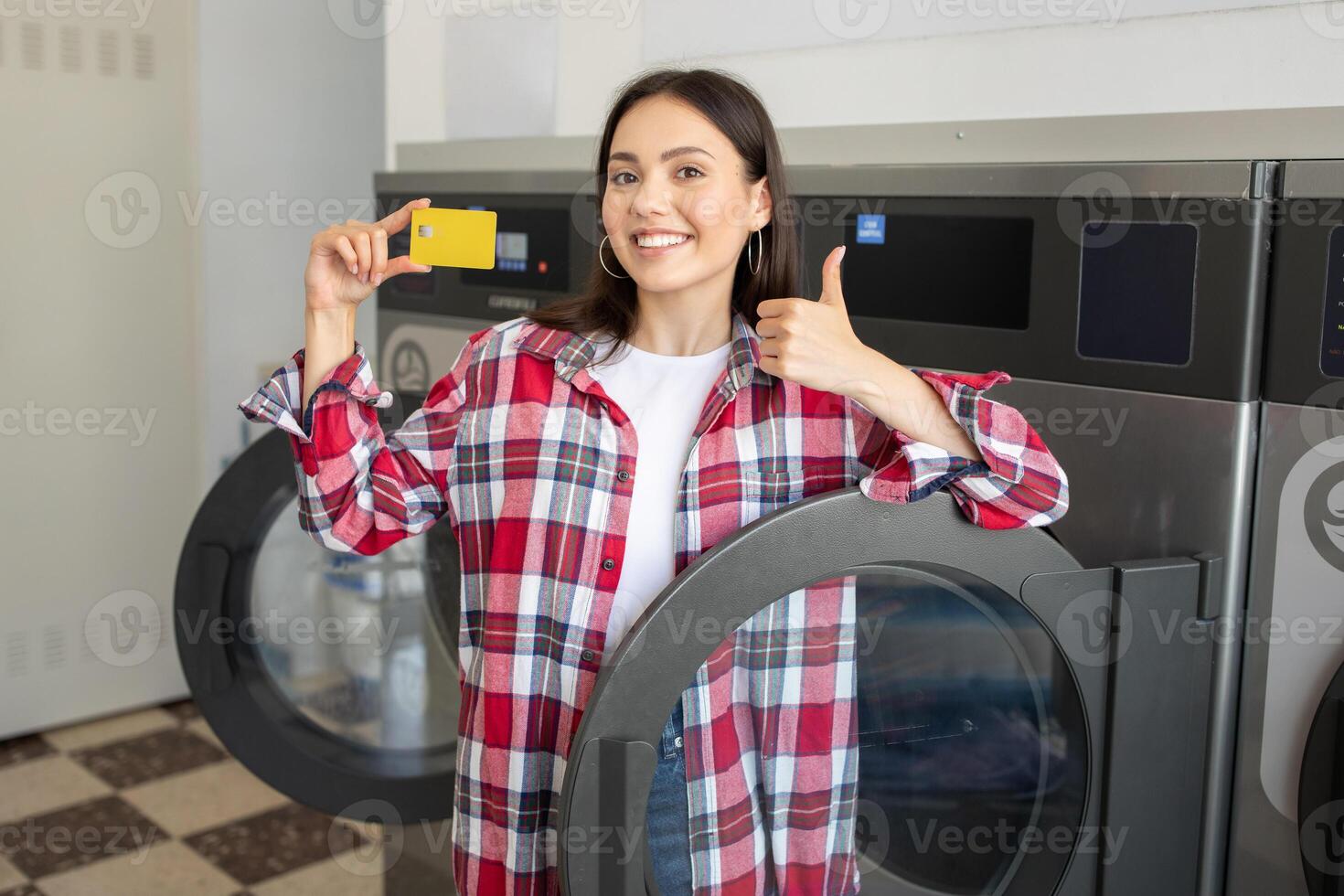 A woman in a red and white plaid shirt is standing in front of a washing machine at a laundromat. She is holding a yellow card in her right hand and giving a thumbs-up with her left hand. photo