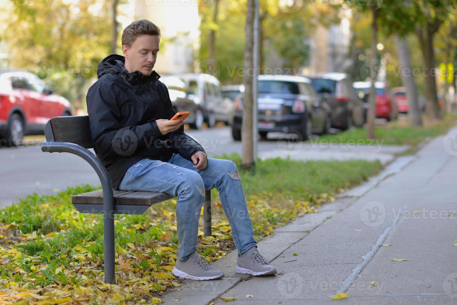 A man chatting texting message using a mobile smartphone device sitting on a bench at the street on a sunny autumn day photo