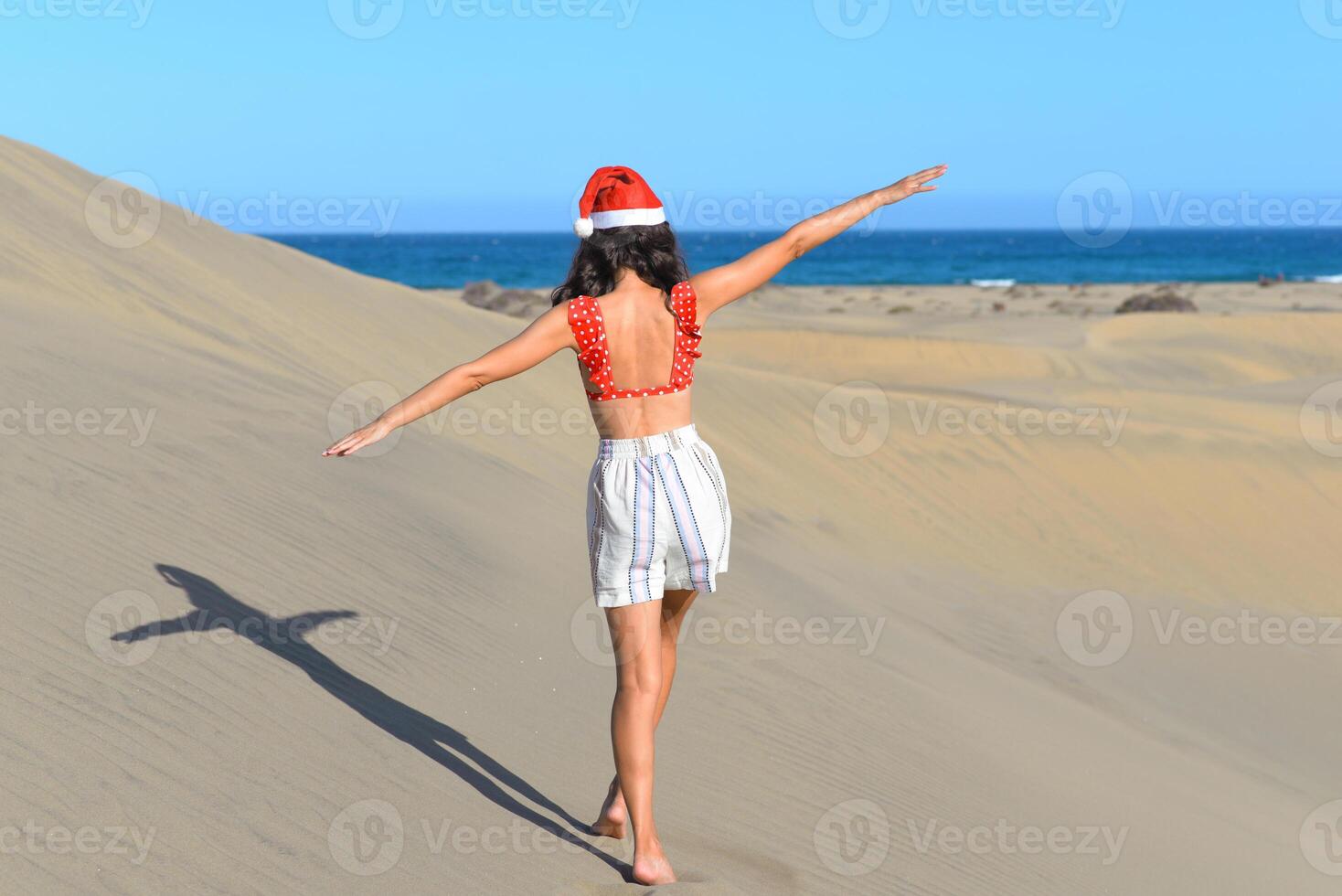 A girl in a dune wearing a Santa hat and a red swimsuit. Back view. The concept of a Christmas vacation in hot countries, at the sea. Dune near the sea in Maspalomas, Gran Canaria photo