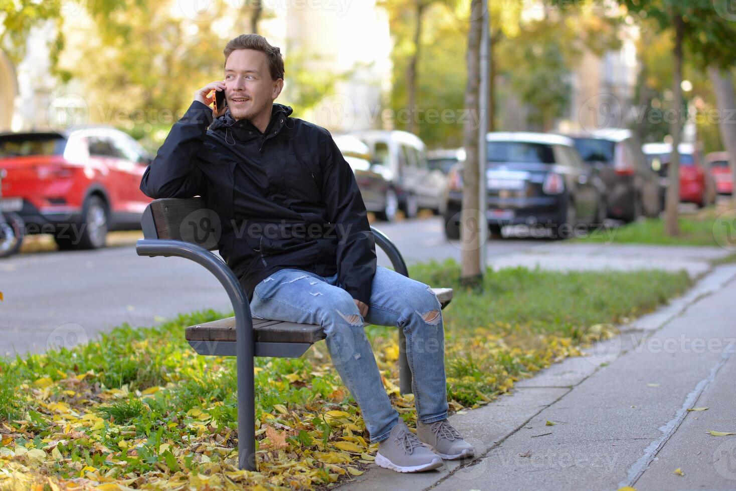A man talking on a phone sitting on a bench at the street on a sunny autumn day. A man uses a mobile smartphone device. Surprise, wonder and happy astonishment emotional expression photo