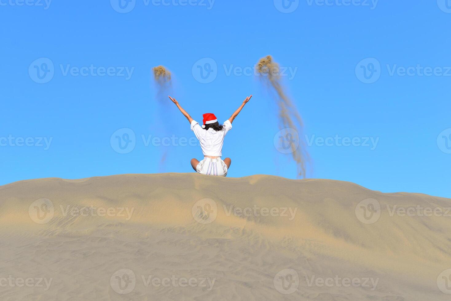 A girl in a dune wearing a Santa hat. A woman throws sand up. Back view. The concept of a Christmas vacation in hot countries, at the sea. Dune near the sea in Maspalomas, Gran Canaria photo