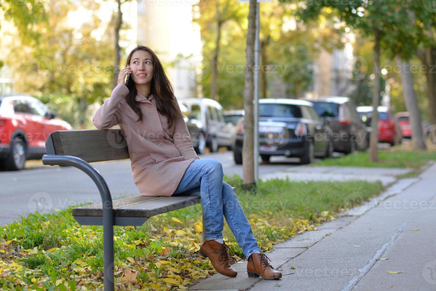 A woman talking on a phone sitting on a bench at the street on a sunny autumn day. A woman smiles while using a mobile smartphone device photo