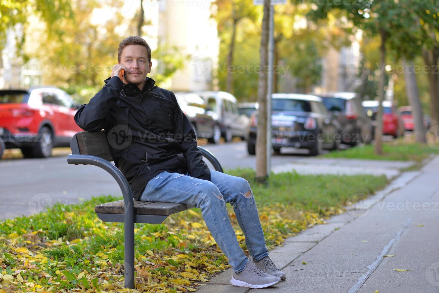 A man talking on a phone sitting on a bench at the street on a sunny autumn day. A man uses a mobile smartphone device photo