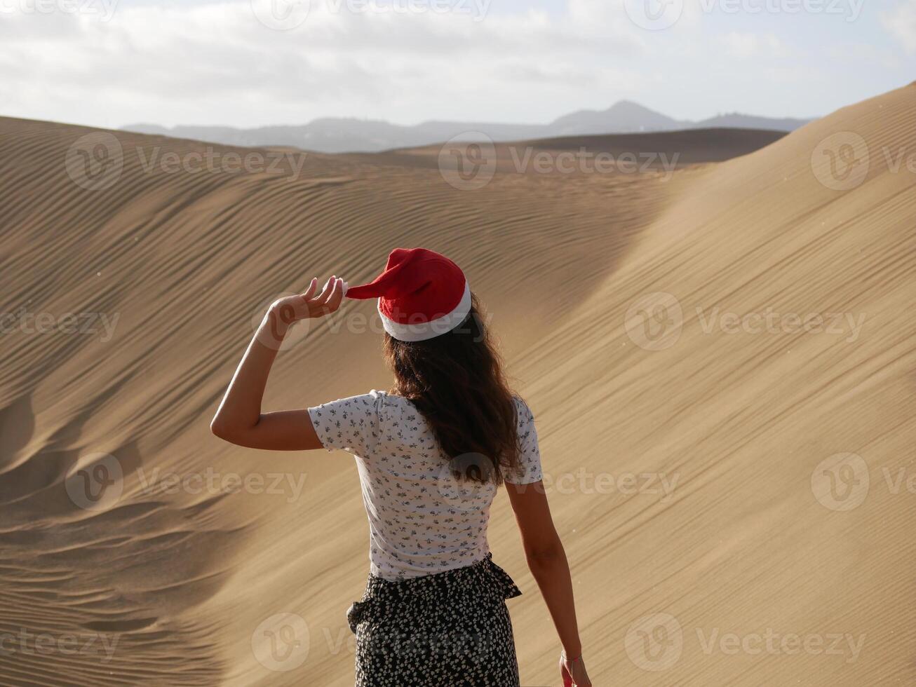 A girl in a dune wearing a Santa hat. Back view. The concept of a Christmas vacation in hot countries, at the sea. Dune near the sea in Maspalomas, Gran Canaria photo