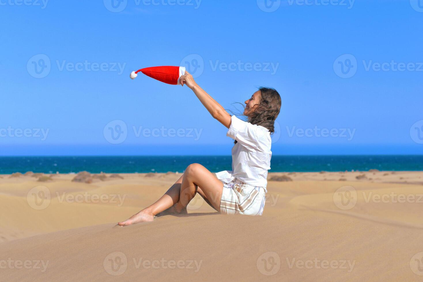 A girl in a dune with a Santa hat fluttering in the wind. Side view. The concept of a Christmas vacation in hot countries, at the sea. Dune near the sea in Maspalomas, Gran Canaria photo