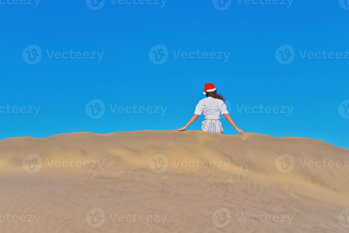 A girl in a dune wearing a Santa hat. Back view. The concept of a Christmas vacation in hot countries, at the sea. Dune near the sea in Maspalomas, Gran Canaria photo