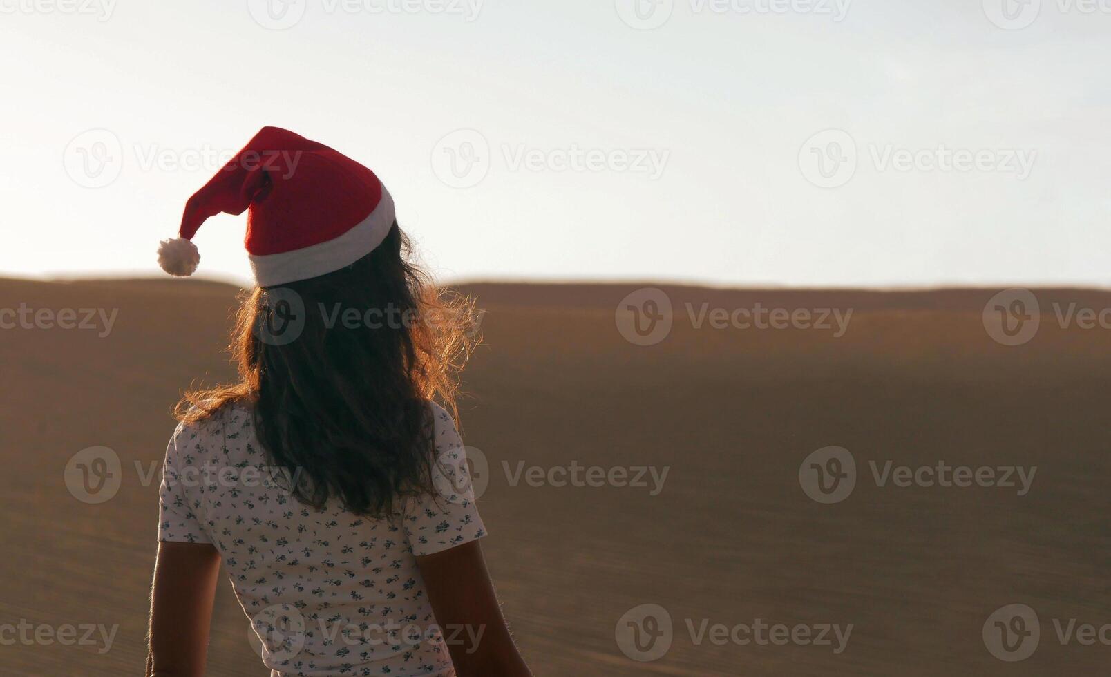 A girl in a dune wearing a Santa hat. Back view. The concept of a Christmas vacation in hot countries, at the sea. Dune near the sea in Maspalomas, Gran Canaria photo