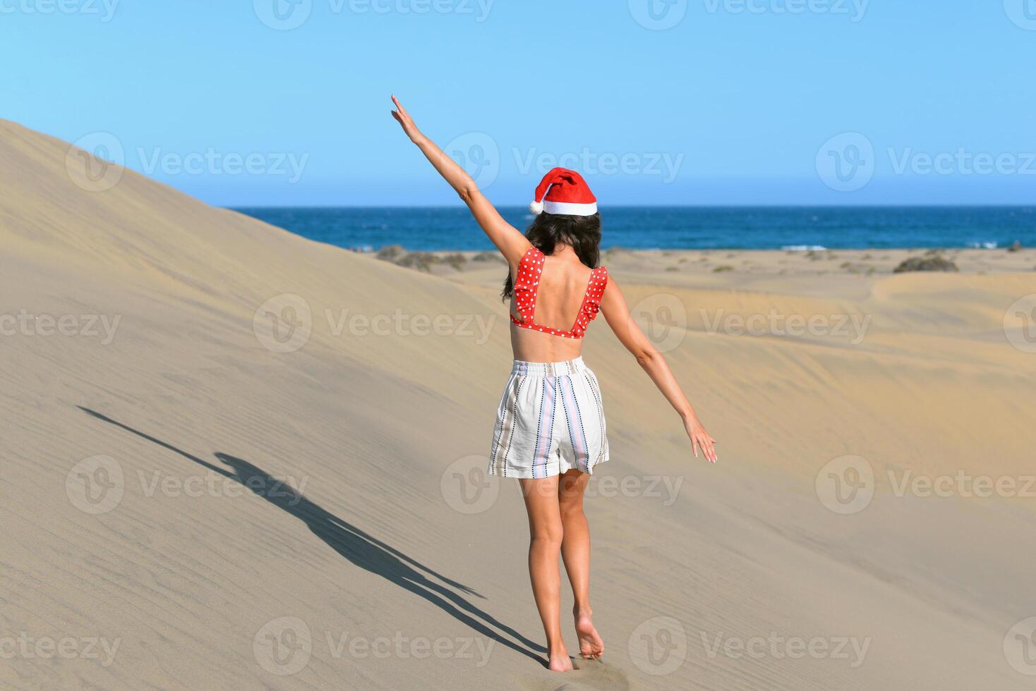 A girl in a dune wearing a Santa hat and a red swimsuit. Back view. The concept of a Christmas vacation in hot countries, at the sea. Dune near the sea in Maspalomas, Gran Canaria photo