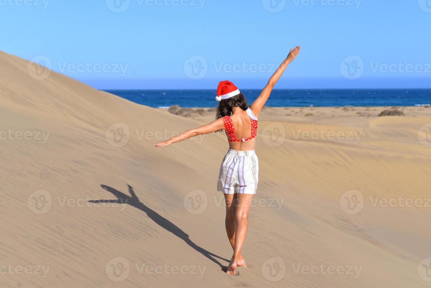 A girl in a dune wearing a Santa hat and a red swimsuit. Back view. The concept of a Christmas vacation in hot countries, at the sea. Dune near the sea in Maspalomas, Gran Canaria photo