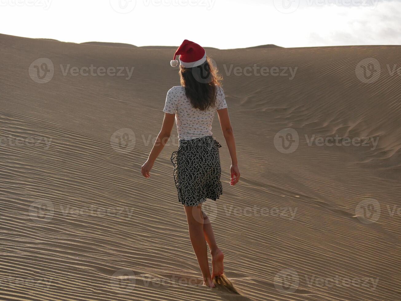 A girl in a dune wearing a Santa hat. Back view. The concept of a Christmas vacation in hot countries, at the sea. Dune near the sea in Maspalomas, Gran Canaria photo