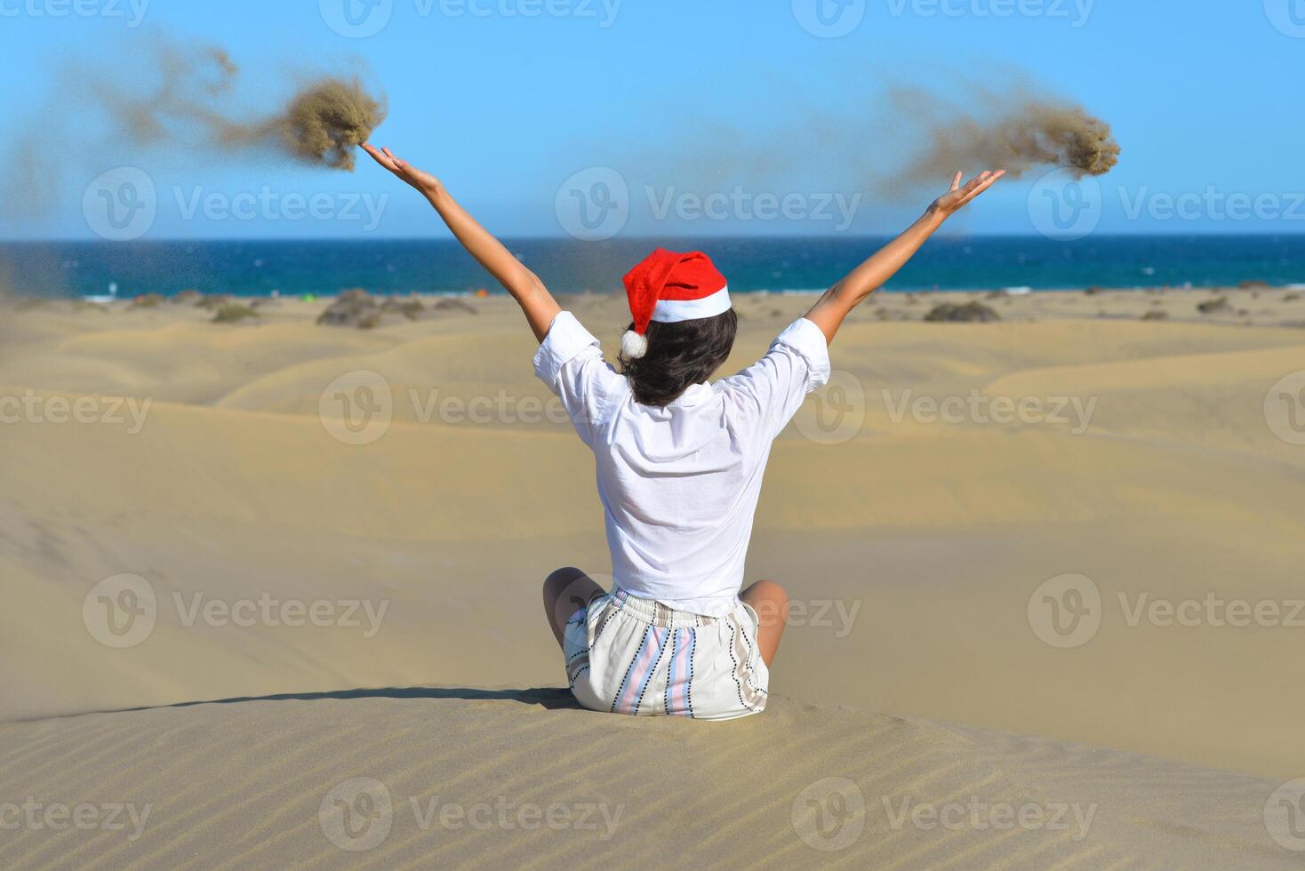 A girl in a dune wearing a Santa hat. A woman throws sand up. Back view. The concept of a Christmas vacation in hot countries, at the sea. Dune near the sea in Maspalomas, Gran Canaria photo