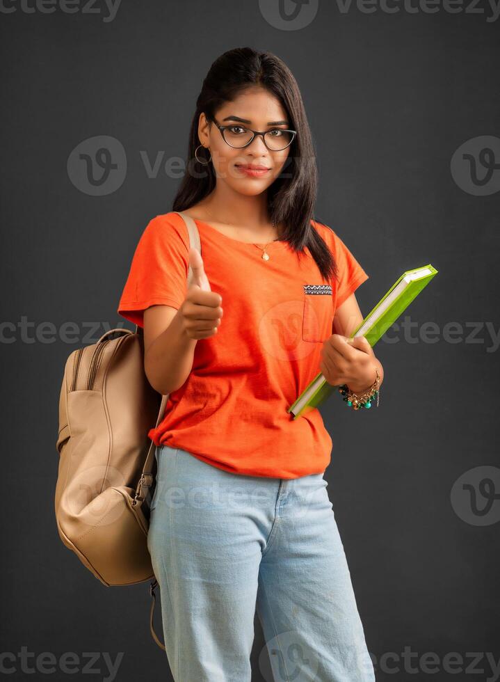 Beautiful young girl with a backpack standing and holding a notebook, posing on a grey background. photo