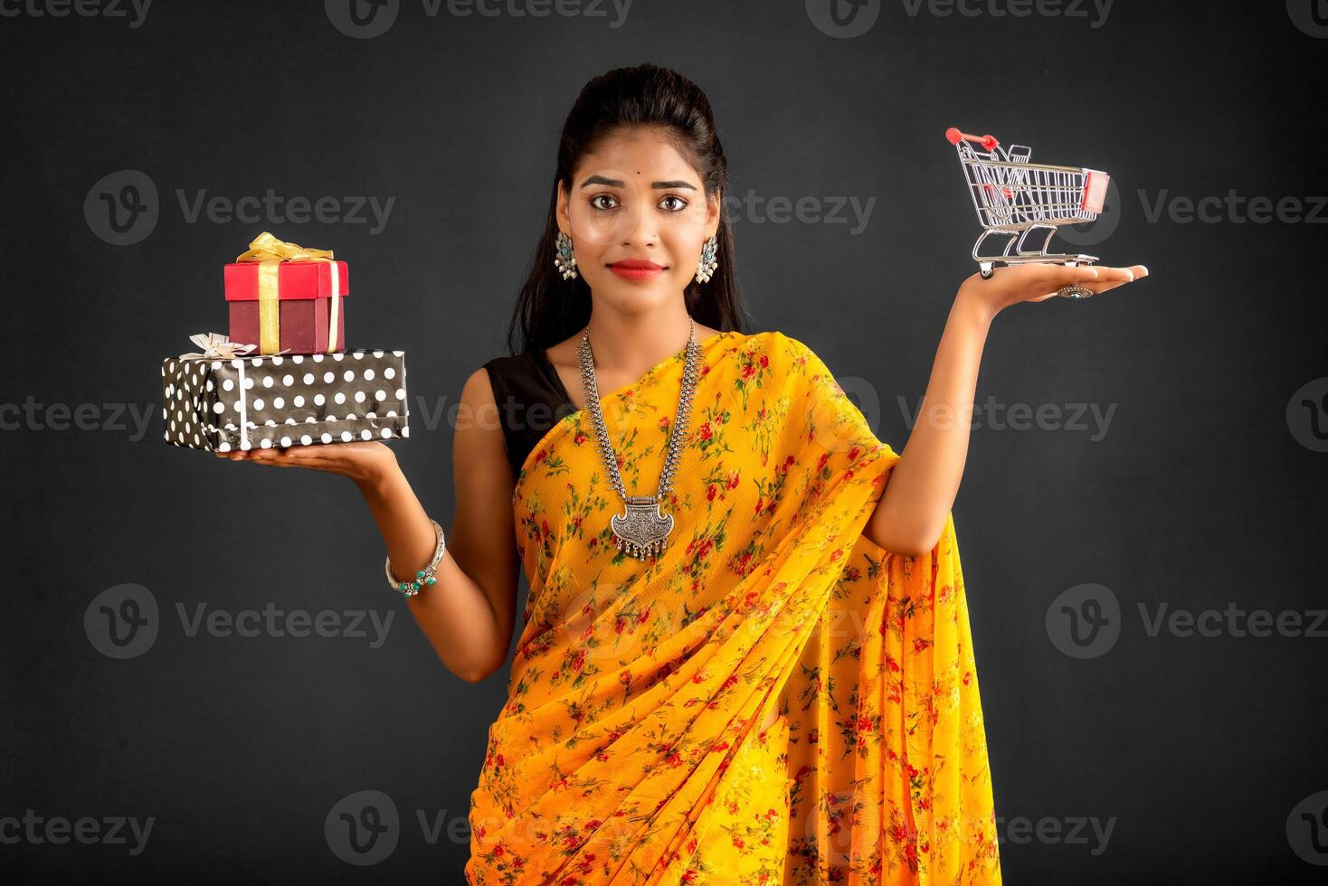 Beautiful young girl or woman holding and posing with a miniature trolley shopping cart and gift box on a gray background photo
