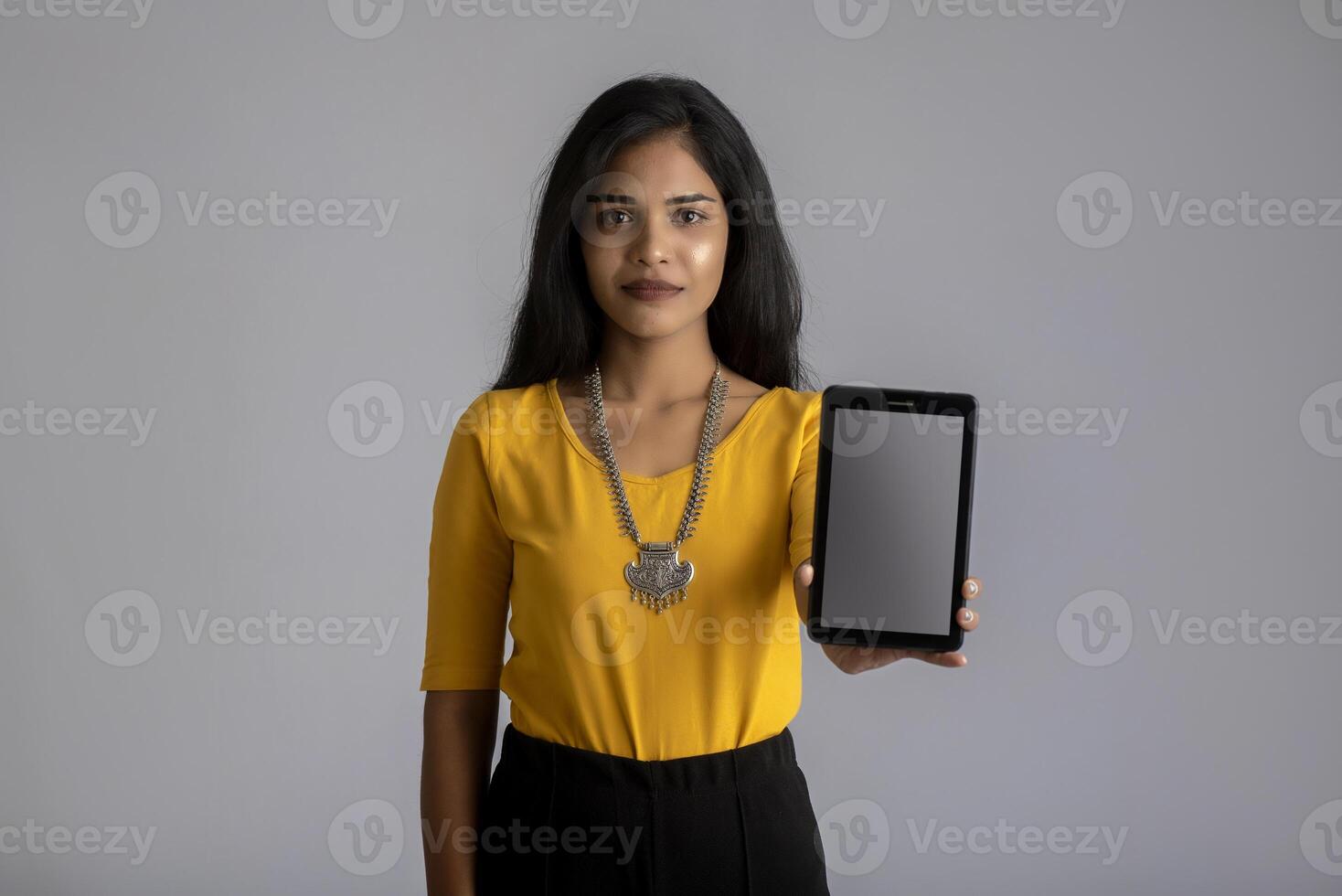 Young beautiful girl holding and showing blank screen of smartphone or mobile or tablet phone on a gray background. photo