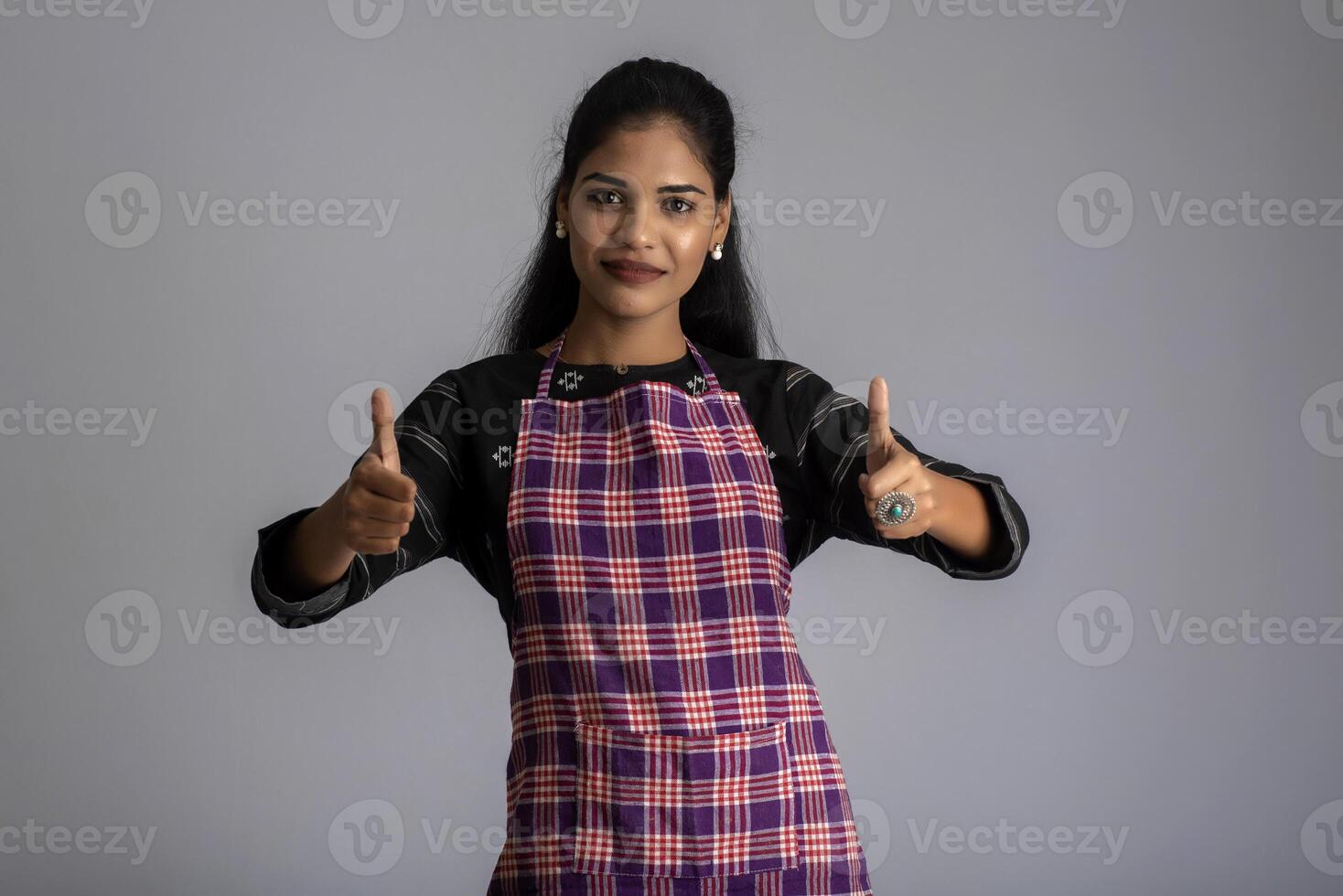 Portrait of Indian woman chef or cook in an apron, presenting, pointing, with ok sign, thumbs up on the grey background. photo
