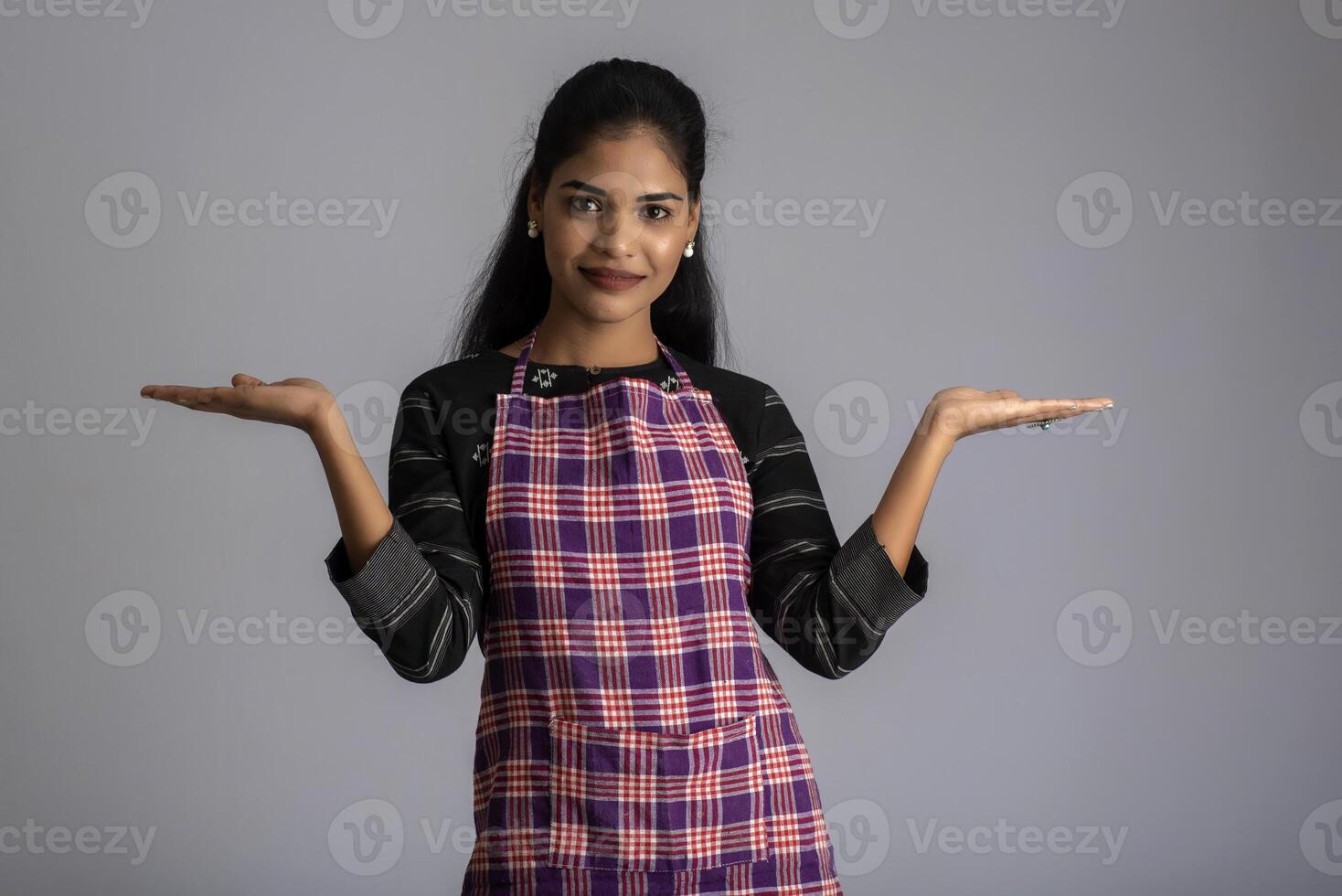 Portrait of Indian woman chef or cook in an apron, presenting, pointing, with ok sign, thumbs up on the grey background. photo