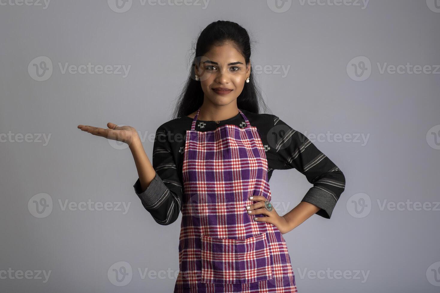 Portrait of Indian woman chef or cook in an apron, presenting, pointing, with ok sign, thumbs up on the grey background. photo