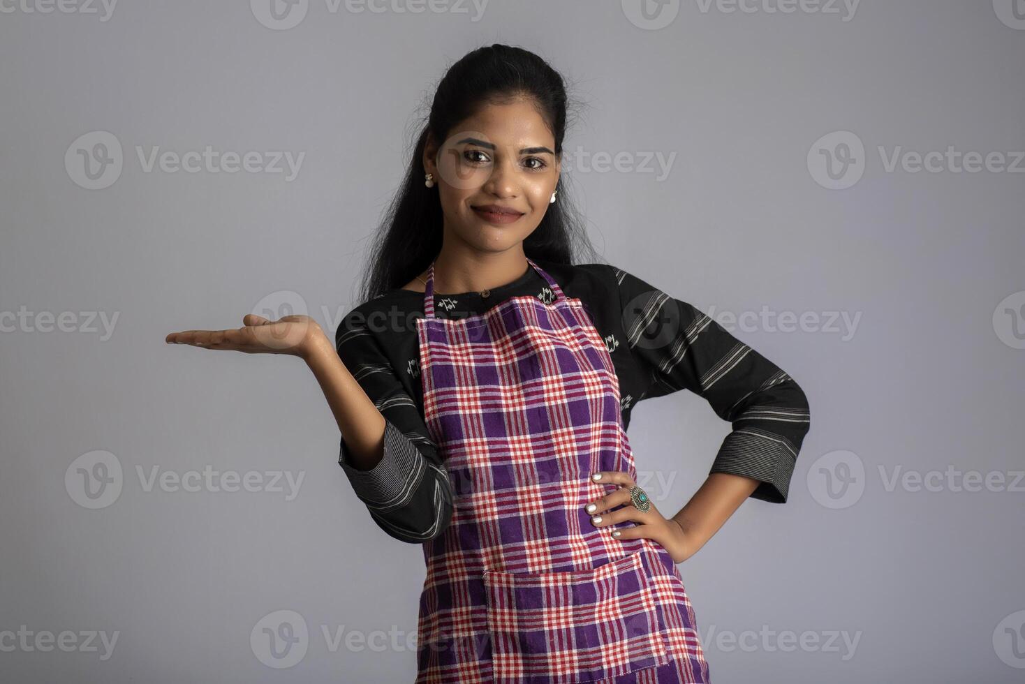 Portrait of Indian woman chef or cook in an apron, presenting, pointing, with ok sign, thumbs up on the grey background. photo