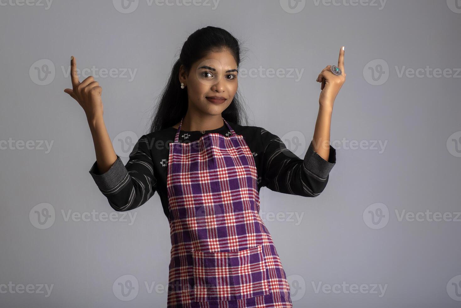 Portrait of Indian woman chef or cook in an apron, presenting, pointing, with ok sign, thumbs up on the grey background. photo